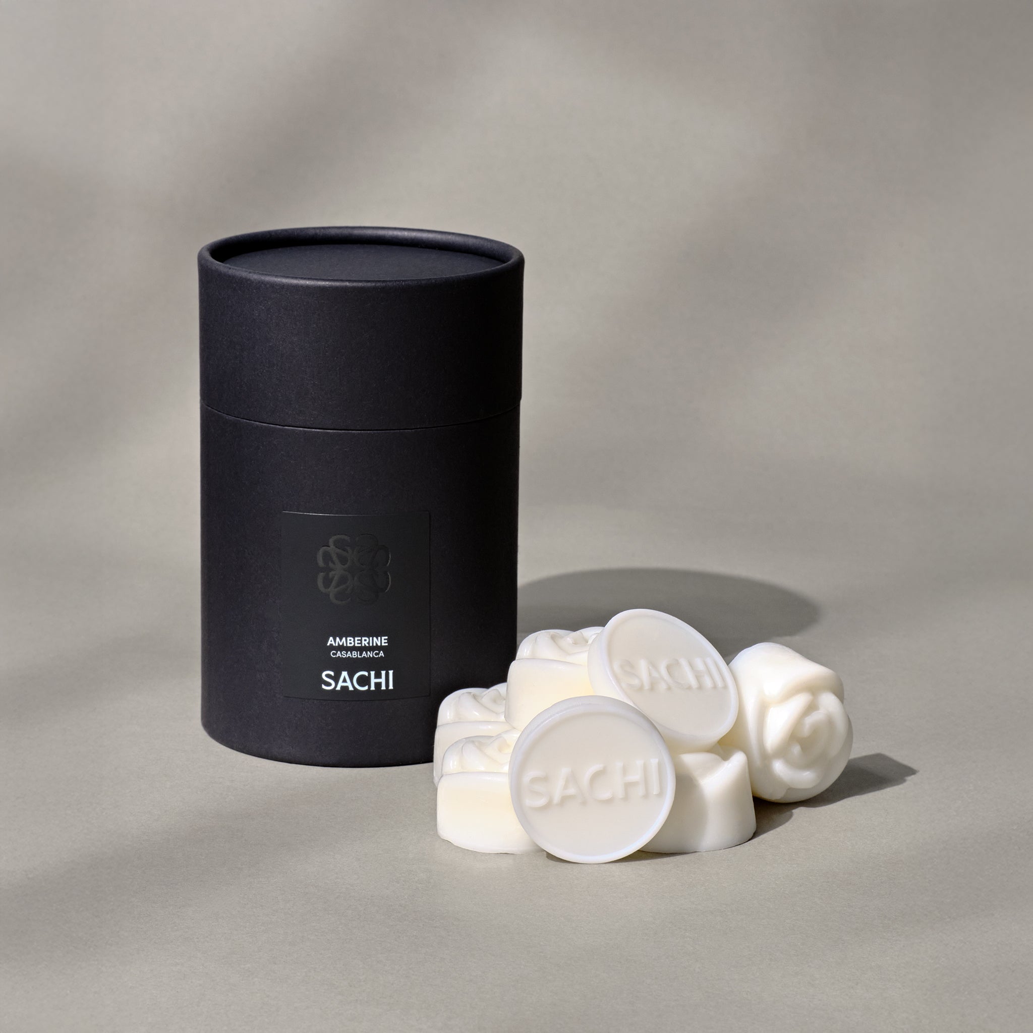 A black cylindrical tube labeled 'SACHI' with multiple round ivory fragrance wax melts in front of it.