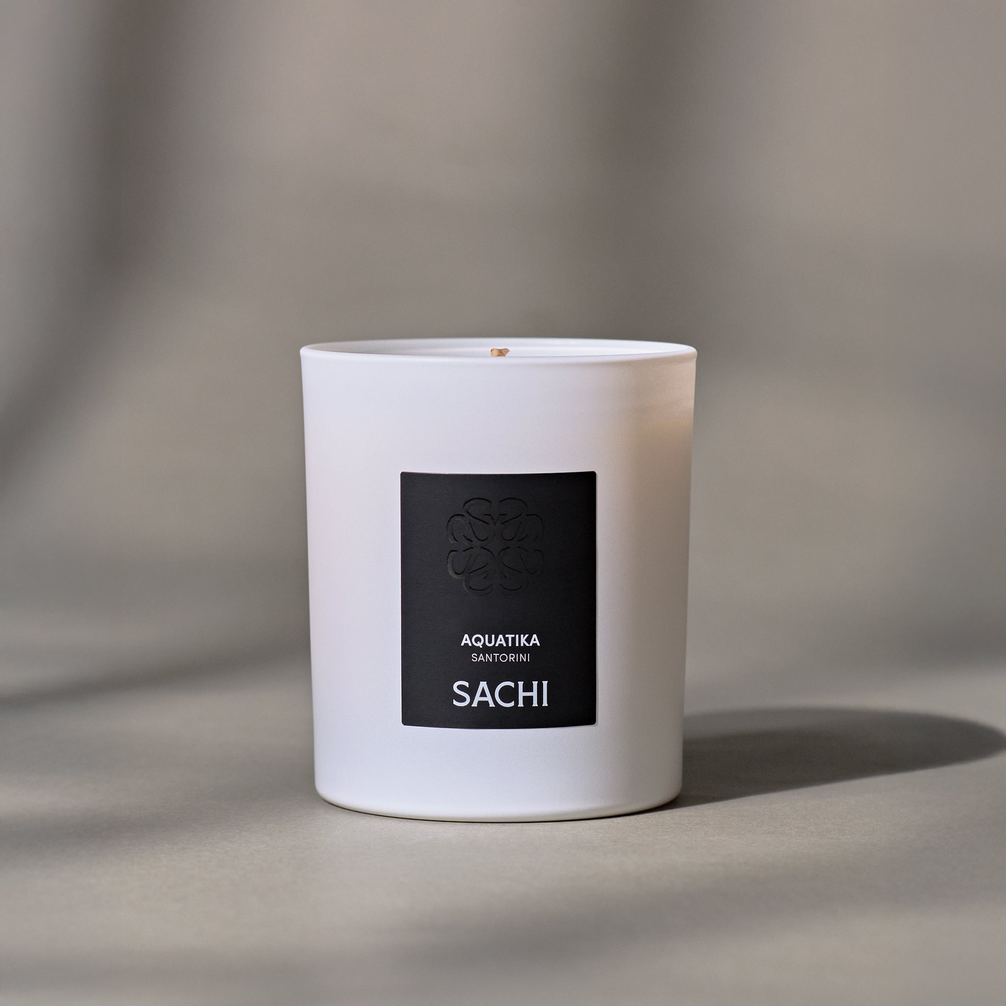 A matte white candle with the brand name 'SACHI' and 'AQUATIKA' on the front. Set on a neutral plain background.