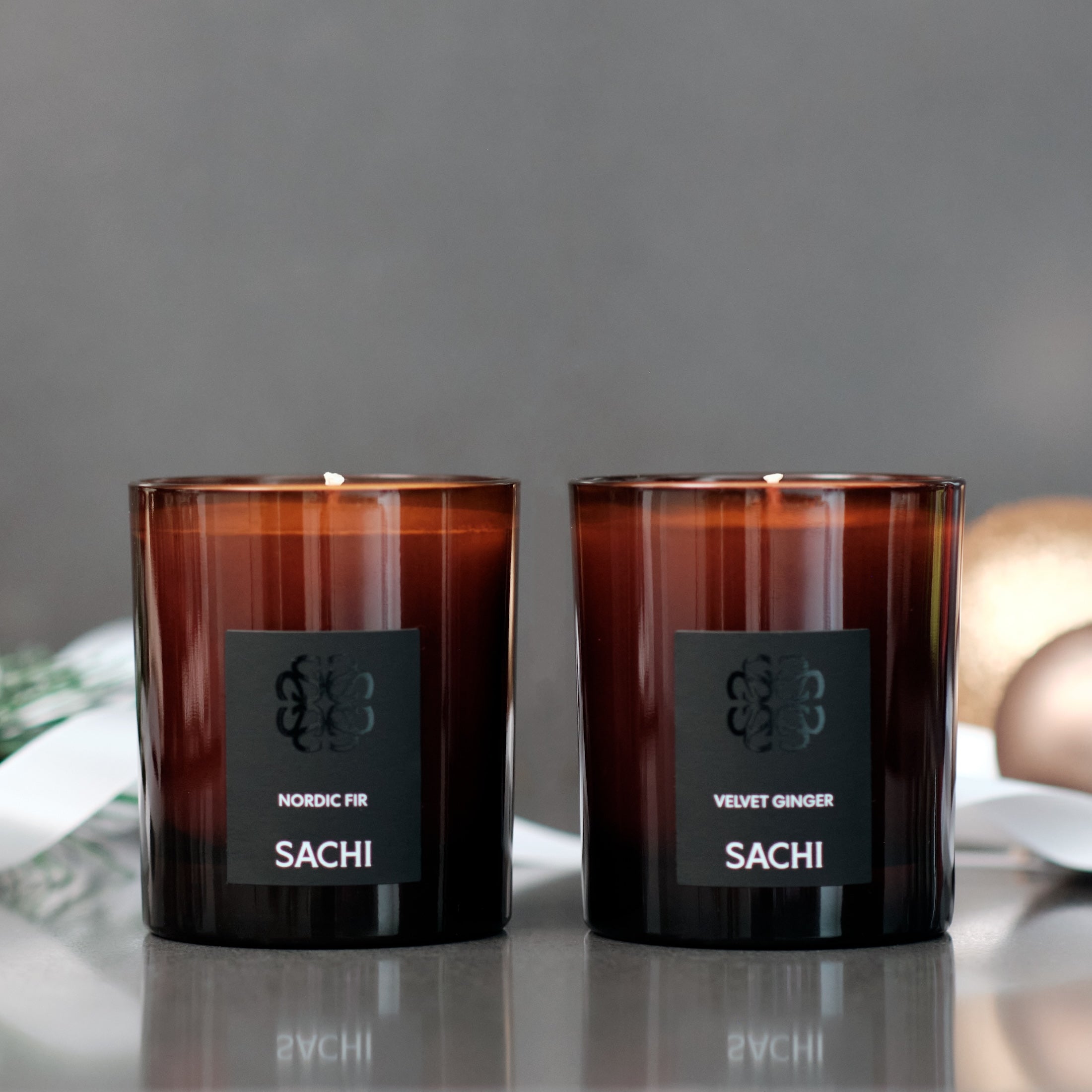 Two SACHI Christmas scented candles with labels on a reflective surface and grey background