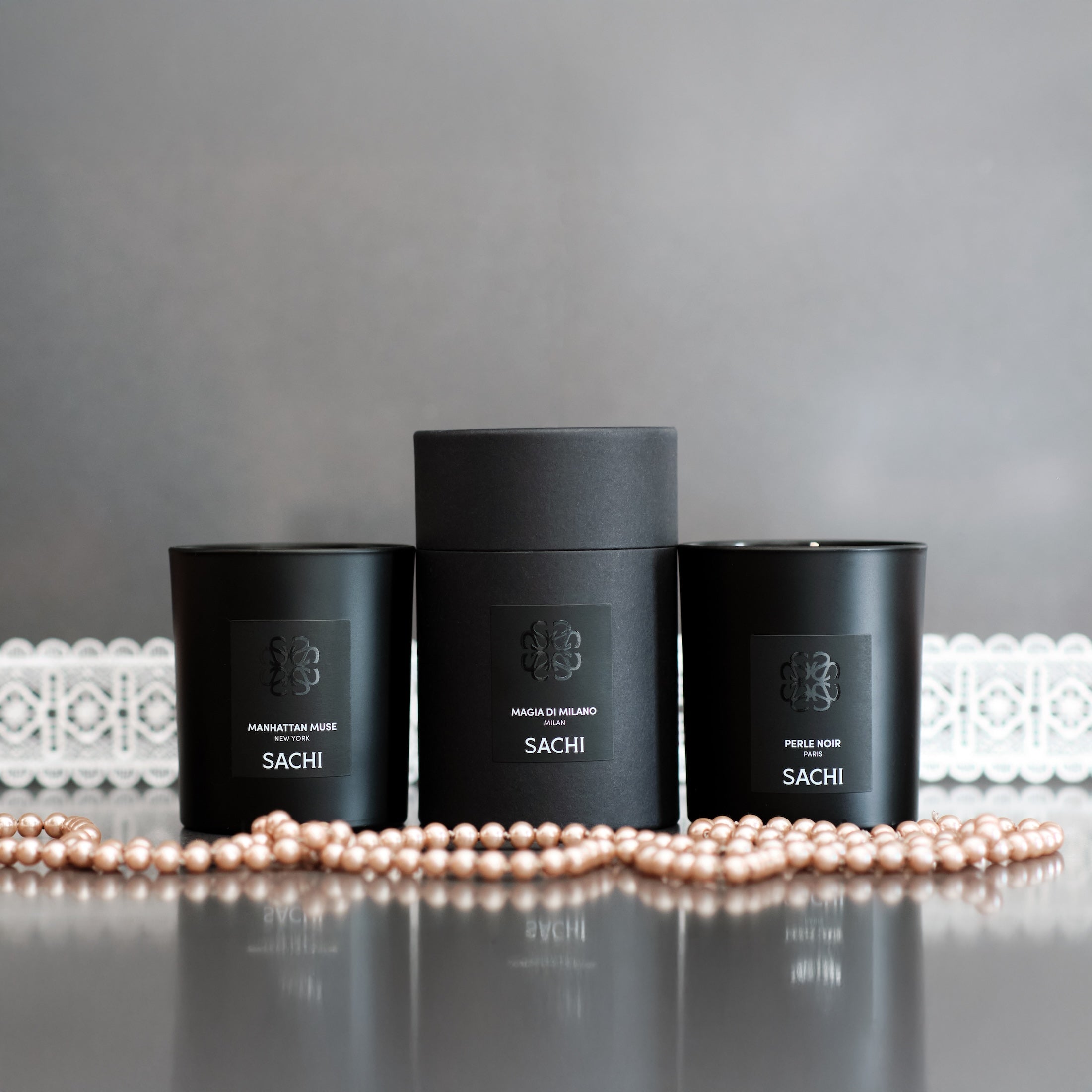 Three black candles labeled 'Sachi' on a reflective surface with pearls in front of a grey background.