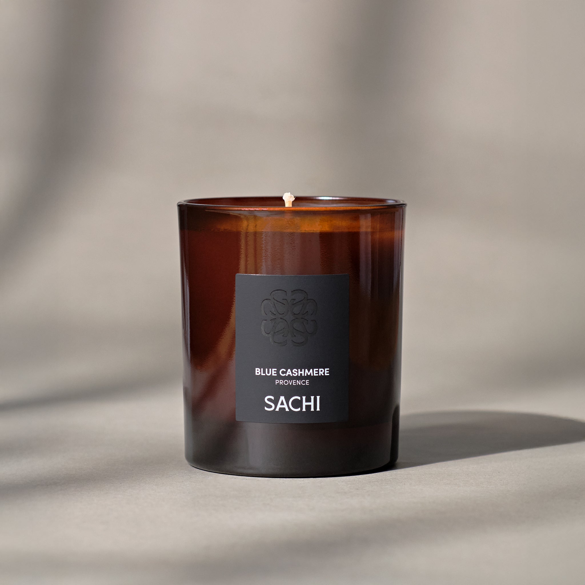 An amber coloured candle with a label that reads 'BLUE CASHMERE and SACHI', set against a neutral background with a shadow cast over the surface.