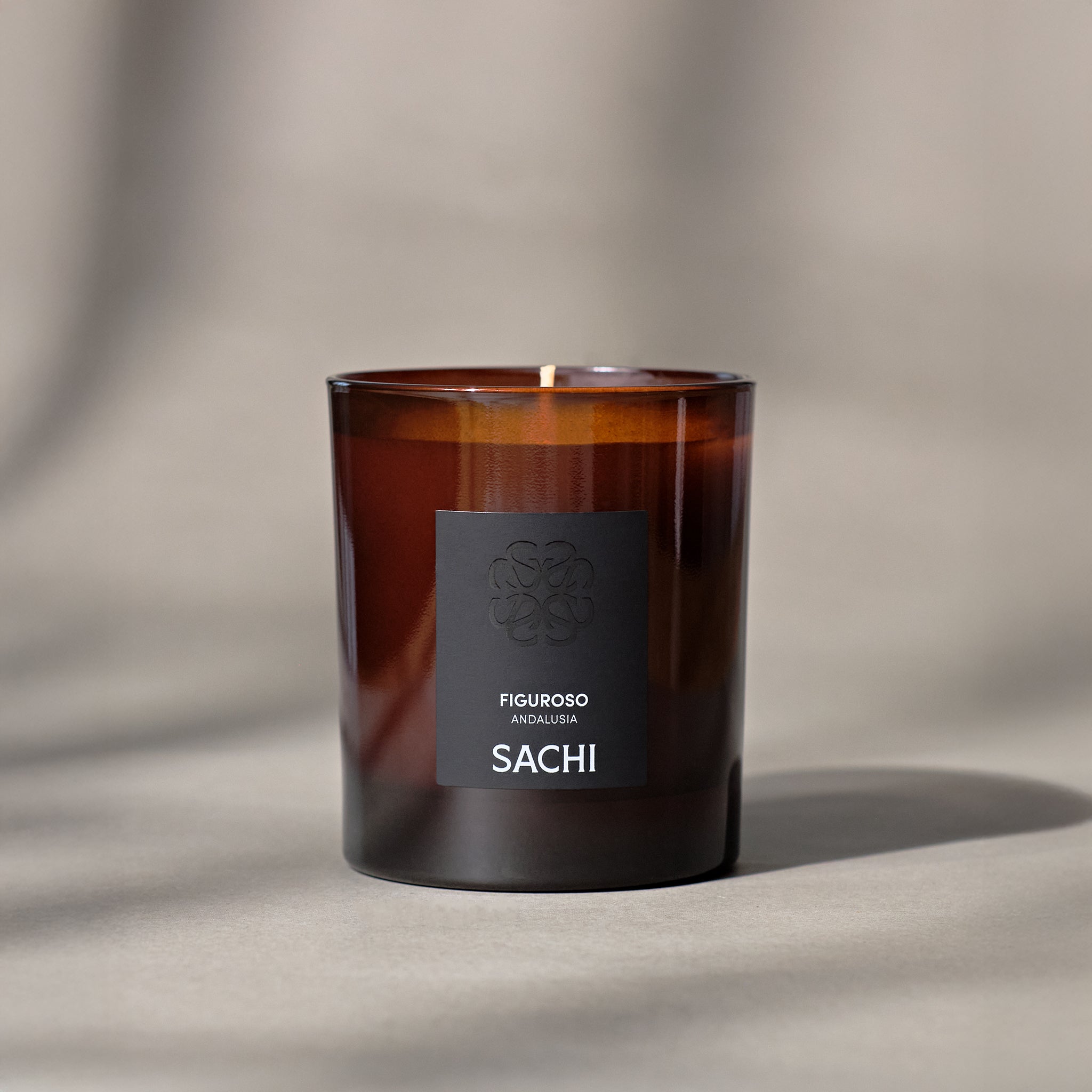 An amber coloured candle with a label that reads 'FIGUROSO and SACHI', set against a neutral background with a shadow cast over the surface.