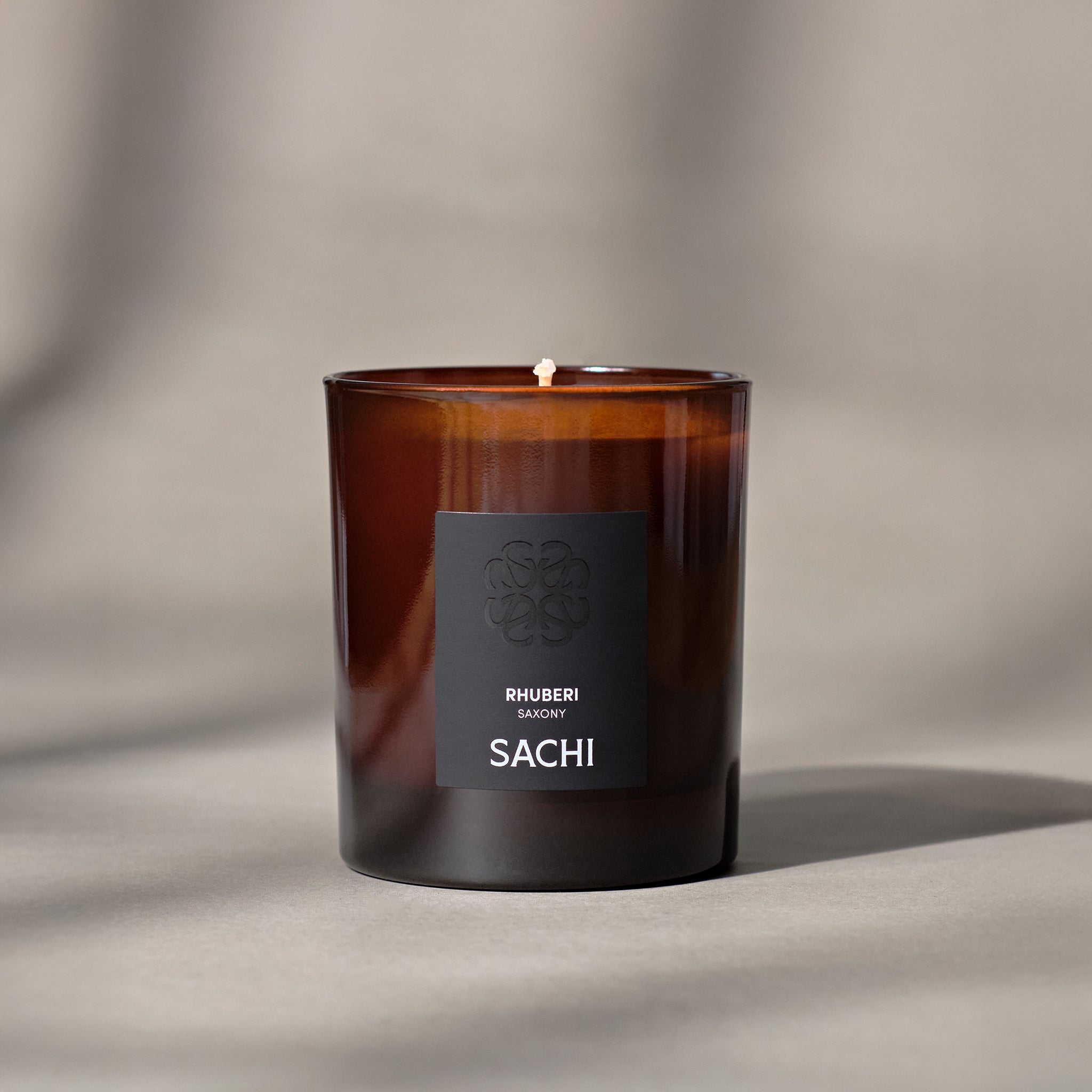 A scented candle with the label 'RHUBERI by SACHI' burning in an amber glass container, with a background of a neutral color.