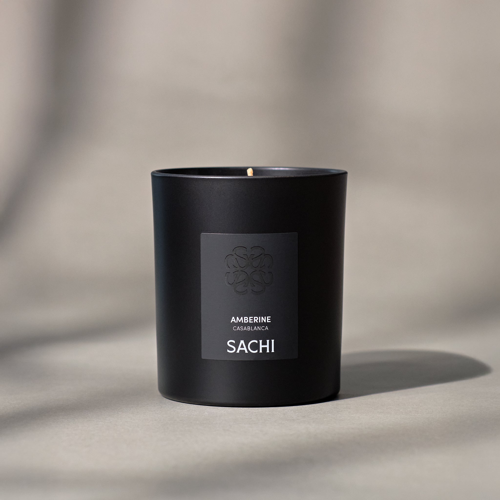 A black candle with a white label that reads 'AMBERINE' and 'SACHI', with a lit wick, placed on a textured surface.