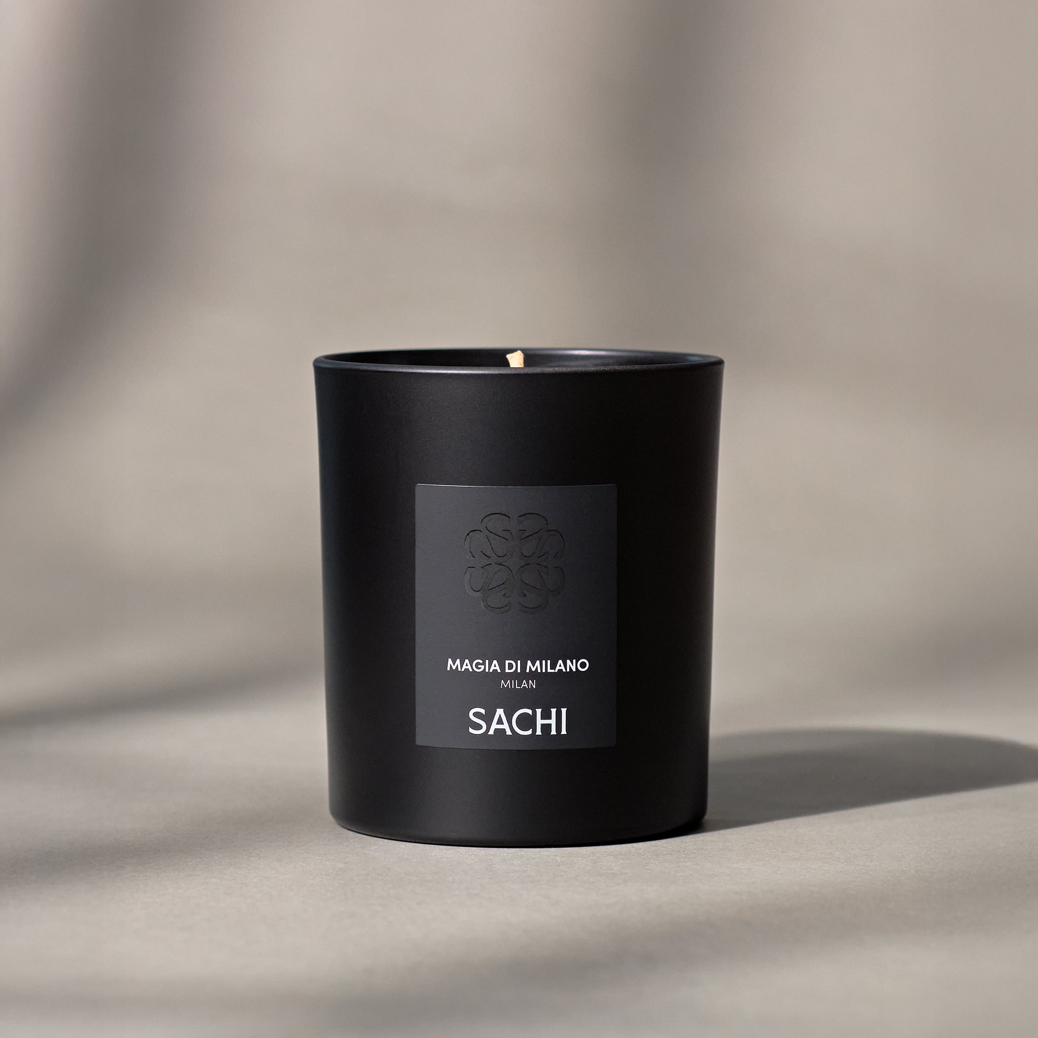 A black candle with the label 'MAGIA DI MILANO' and 'SACHI' on it, placed on a light-colored surface.