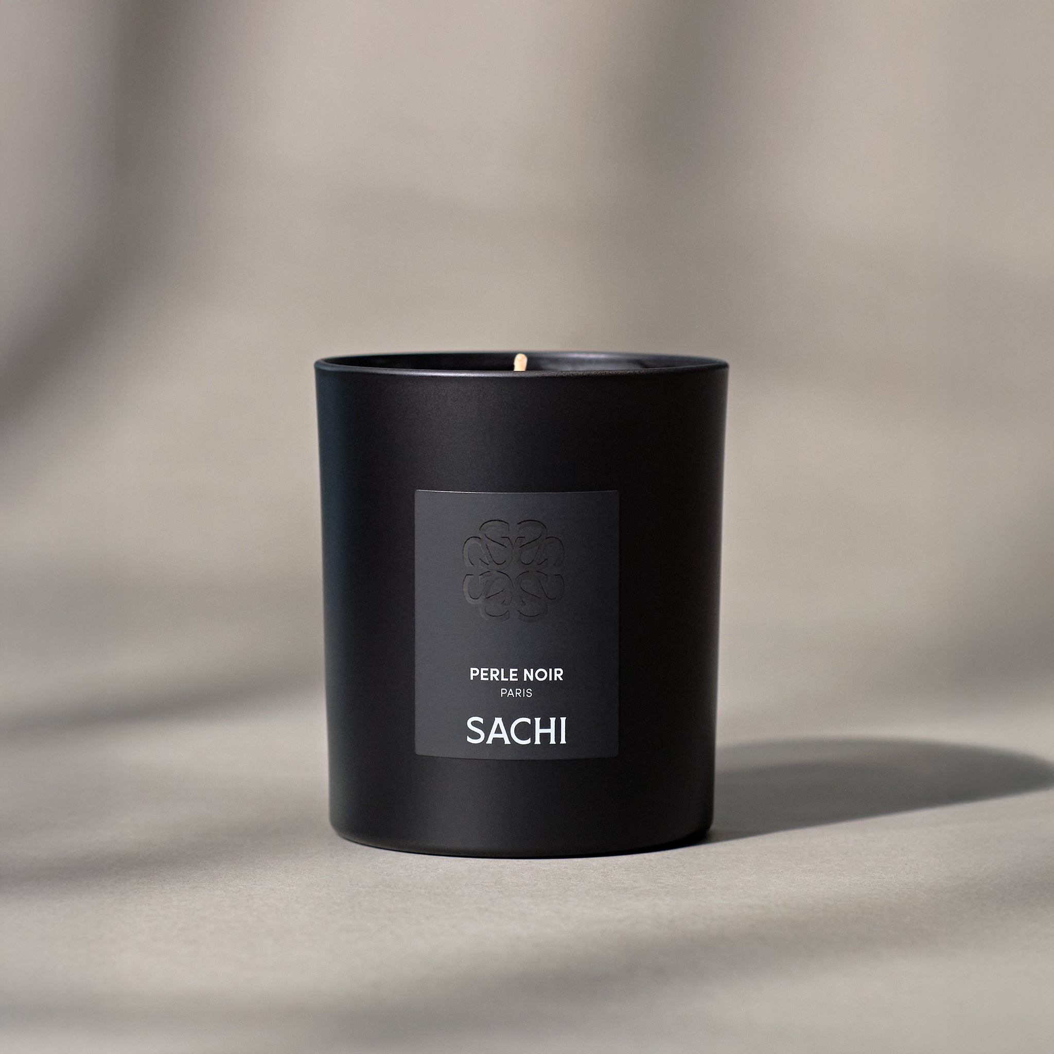 A black candle with the name 'PERLE NOIR' and the brand 'SACHI' printed on a label on the front. On a neutral background.