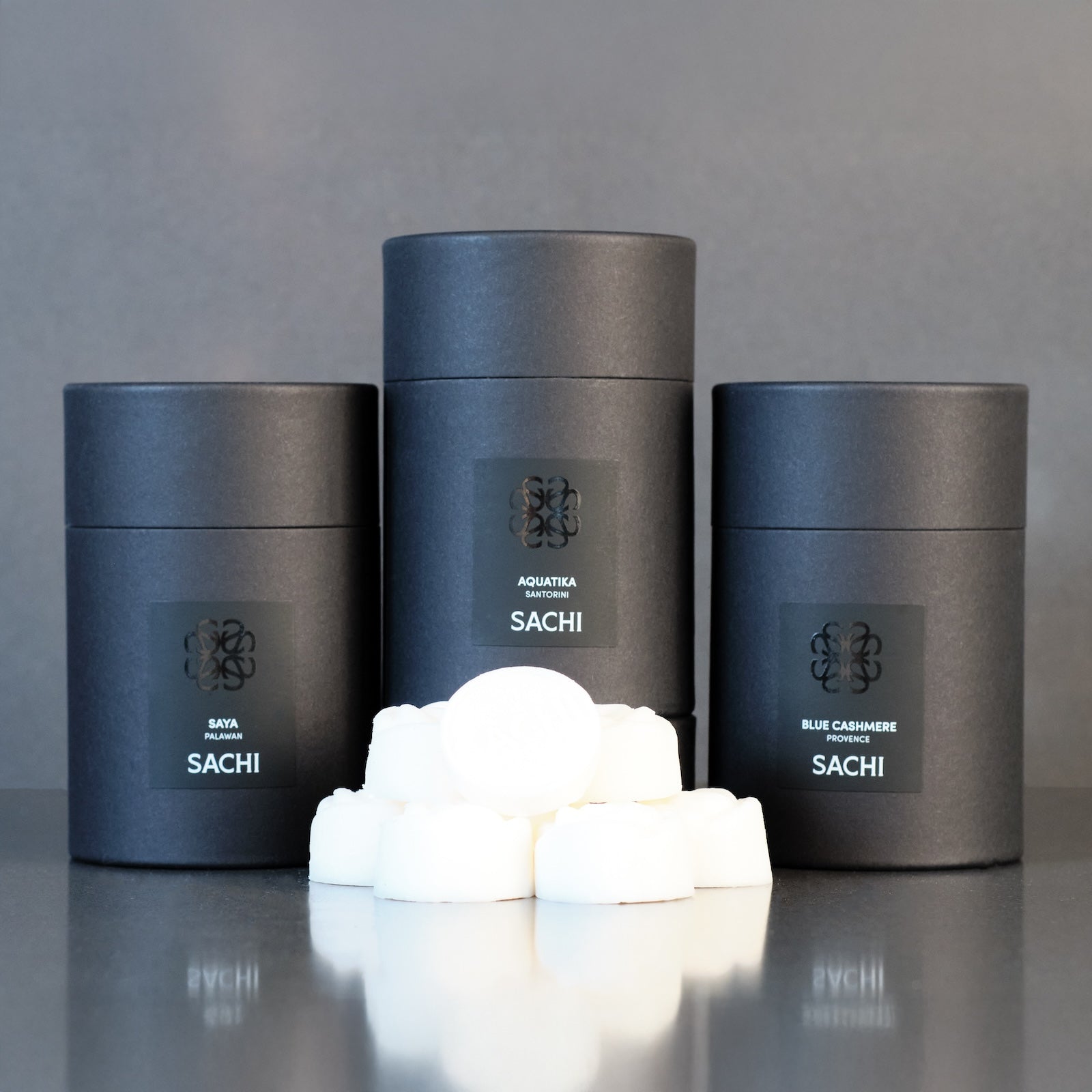 Three black cylindrical containers with 'Sachi' branding on a grey background and wax melts