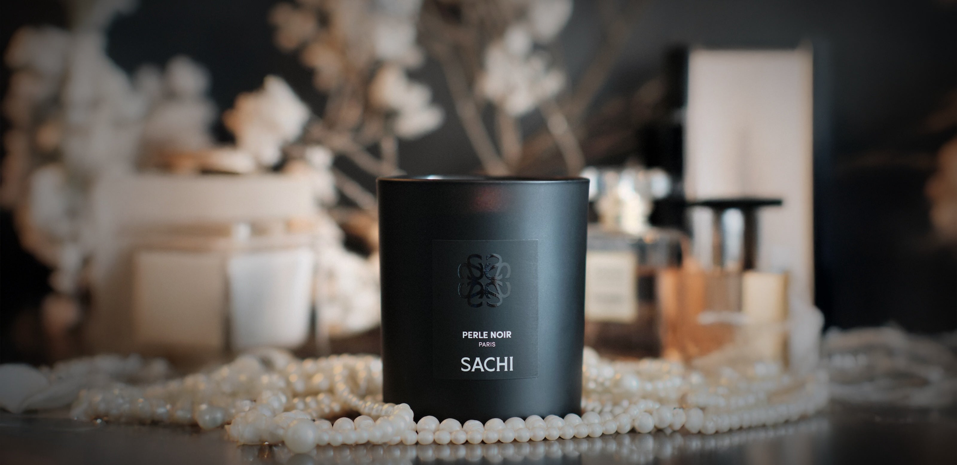 Black Sachi candle named Perle Noir surrounded by pearls with luxury fragrances blurred in the background