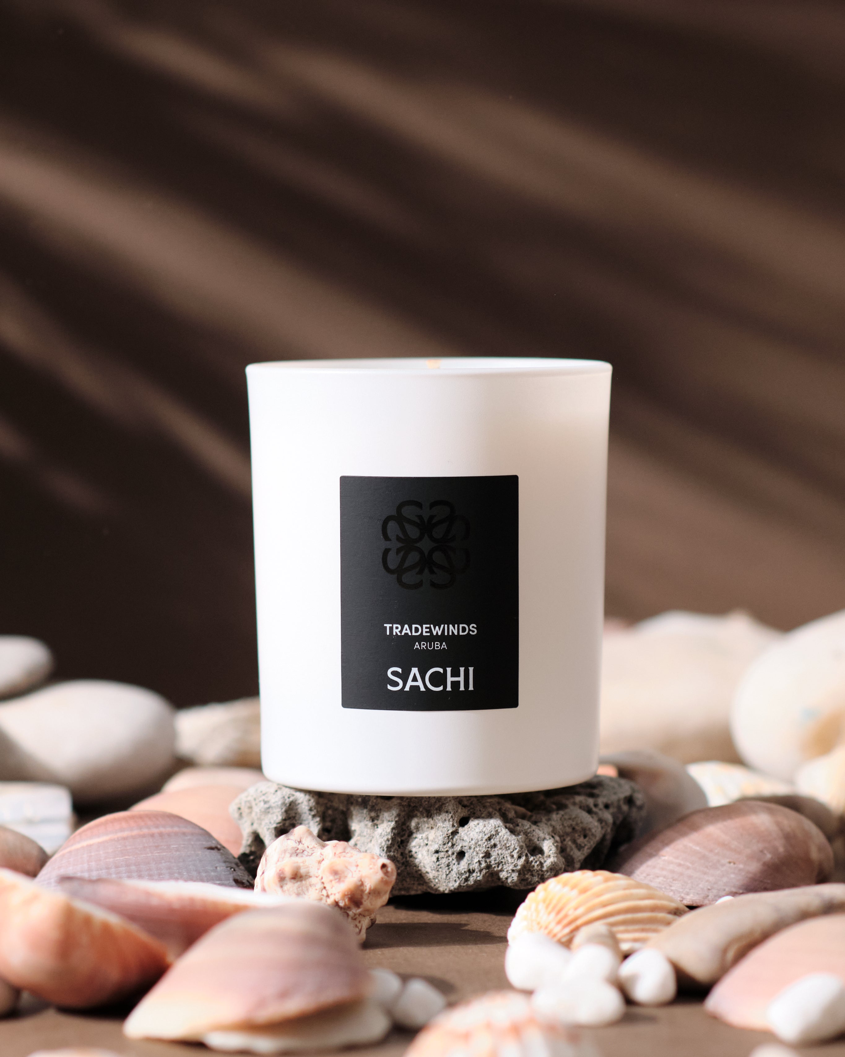 A white SACHI branded candle called TRADEWINDS sitting on a rock surrounded by seashells and the shadow of a palm tree in the background.